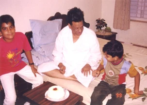 Ajay Rungta (Dadu) sits on a bed with his two grandsons, Bhaiya and Aman. A small cake is on a table in front of them.