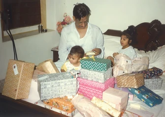 Dadu (Ajay Rungta) on a bed with two children, Chinu and Bhaiya, opening gifts from a large pile of presents surrounding them.