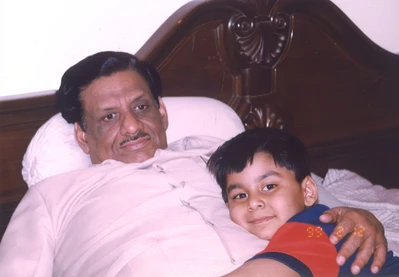 Ajay Rungta (Dadu) lying in bed, smiling gently, with his young grandson Aman resting on his chest. Dadu has his arm around Aman.