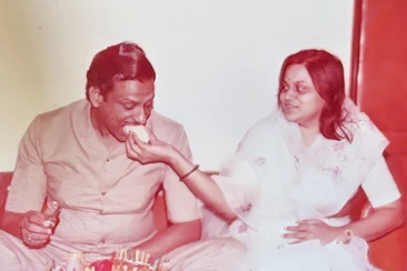 A vintage photograph showing a woman, Ma, gently feeding a piece of cake to a man, Dadu (Ajay Rungta), who is seated beside her.