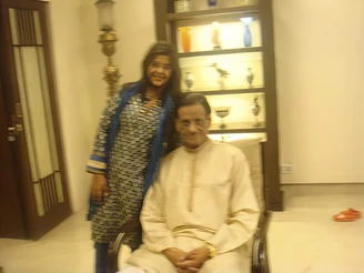 Slightly blurry indoor photo: Ajay Rungta (Dadu) seated, Shivi standing beside him, both smiling. Warm family moment.