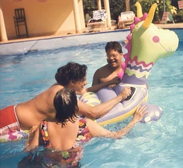 Dadu (Ajay Rungta) smiles while in a swimming pool with two other people and a large, whimsical inflatable water toy.