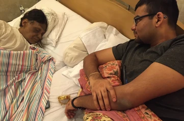 Ajay Rungta (Dadu) asleep in bed, covered with a striped blanket, with his grandson Aman lying awake beside him in a softly lit room.
