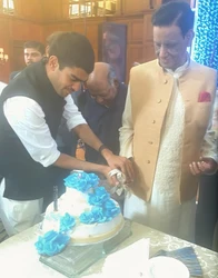 Ajay 'Dadu' Rungta, smiling, and a younger man, Prateek, cutting a three-tiered white cake with blue rose decorations.