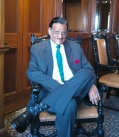 Ajay Rungta (Dadu) seated in an ornate chair, looking dapper in a grey suit with green tie and red pocket square, smiling warmly.