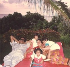 Ajay Rungta (Dadu) relaxing on a red lounge chair outdoors with his wife and three granddaughters. Palm fronds visible.