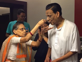 An elderly woman in a traditional saree feeds Dadu Ajay Rungta from a spoon while two others look on in a home setting.