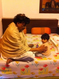 Ajay Rungta (Dadu) in a shawl, sitting on a floral bedsheet, gently holding his young grandchild Ishu. A warm, loving interaction.