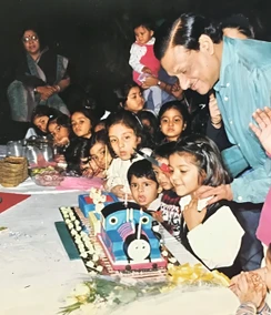Dadu (Ajay Rungta) at a kids' birthday party, looking at a Thomas the Tank Engine cake with many children gathered around.