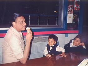 Ajay Rungta, known as Dadu, smiles while eating an ice cream cone, seated with his two young grandsons at a diner table.