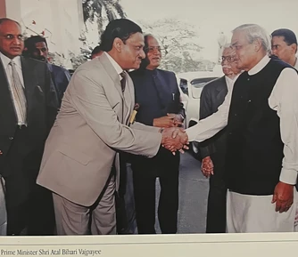 Dadu (Ajay Rungta) in a suit shakes hands with former PM Atal Bihari Vajpayee, dressed in a kurta and vest, outdoors.