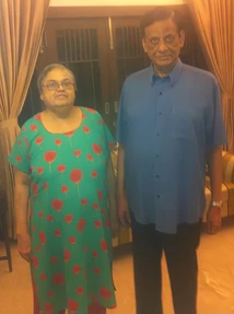 A photograph of Ajay Rungta (Dadu) in a blue shirt and Ma in a green patterned top, standing side-by-side indoors.