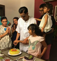 Dadu (Ajay Rungta) stands with his young granddaughter, both holding a knife to cut a colorful fruit cake. Family members watch and smile.