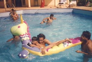 Ajay Rungta (Dadu) and family members in a sunlit swimming pool, with Dadu and a young child on a large colorful inflatable unicorn. Others swim nearby.