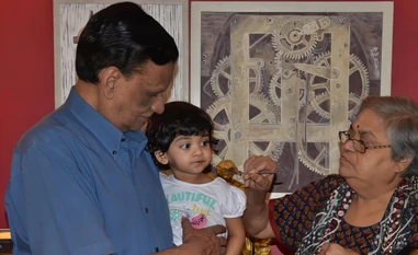 Ajay Rungta (Dadu) holds a young grandchild while a woman offers the child food with a spoon. Indoor family scene.