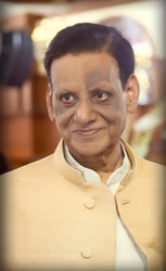 Close-up portrait of Dadu (Ajay Rungta), an elderly Indian man, smiling. He wears a cream Nehru jacket. Warm, blurred background.