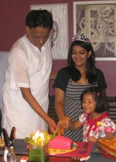 Dadu (Ajay Rungta) and his two granddaughters cutting a pink birthday cake. The older girl wears a tiara, all are smiling.