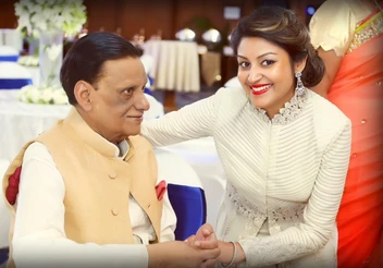 Ajay Rungta (Dadu) in a beige waistcoat, seated and holding hands with Chinu, who wears an elegant white dress. Both are smiling warmly at an event.
