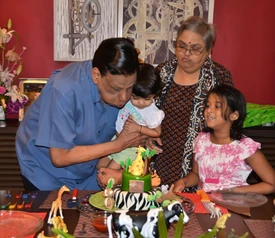 Ajay Rungta (Dadu) lovingly kisses a grandchild by a birthday cake, with family watching. The cake has animal decorations.