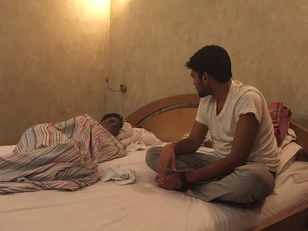Ajay Rungta (Dadu) sleeping under a striped blanket, as Bhaiya, a young man, sits watchfully by his bedside in a dimly lit room.