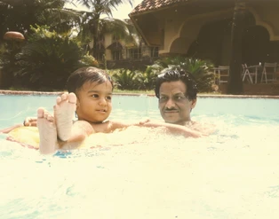 Ajay Rungta (Dadu) smiling in a swimming pool, holding a young child (Bhaiya) who is on a yellow inflatable ring.