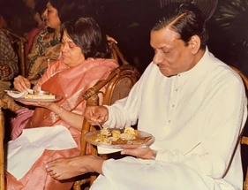 Ajay Rungta (Dadu) and his wife (Ma) are seated side-by-side, each holding a plate and enjoying a meal together indoors.
